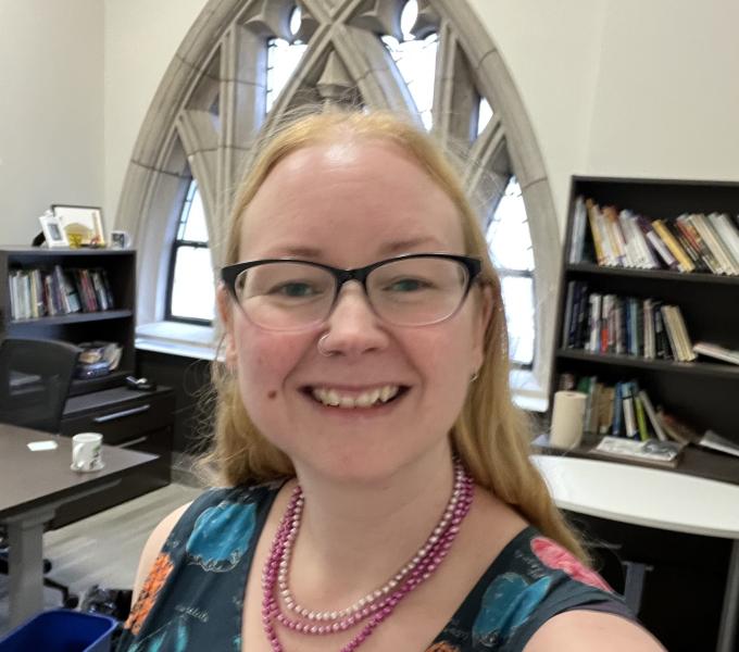 A woman stands in an office in the Cathedral of Learning. She has long hair and is wearing glasses, a pink pearl necklace, and a blue dress. You can see a Neo-gothic window behind her.